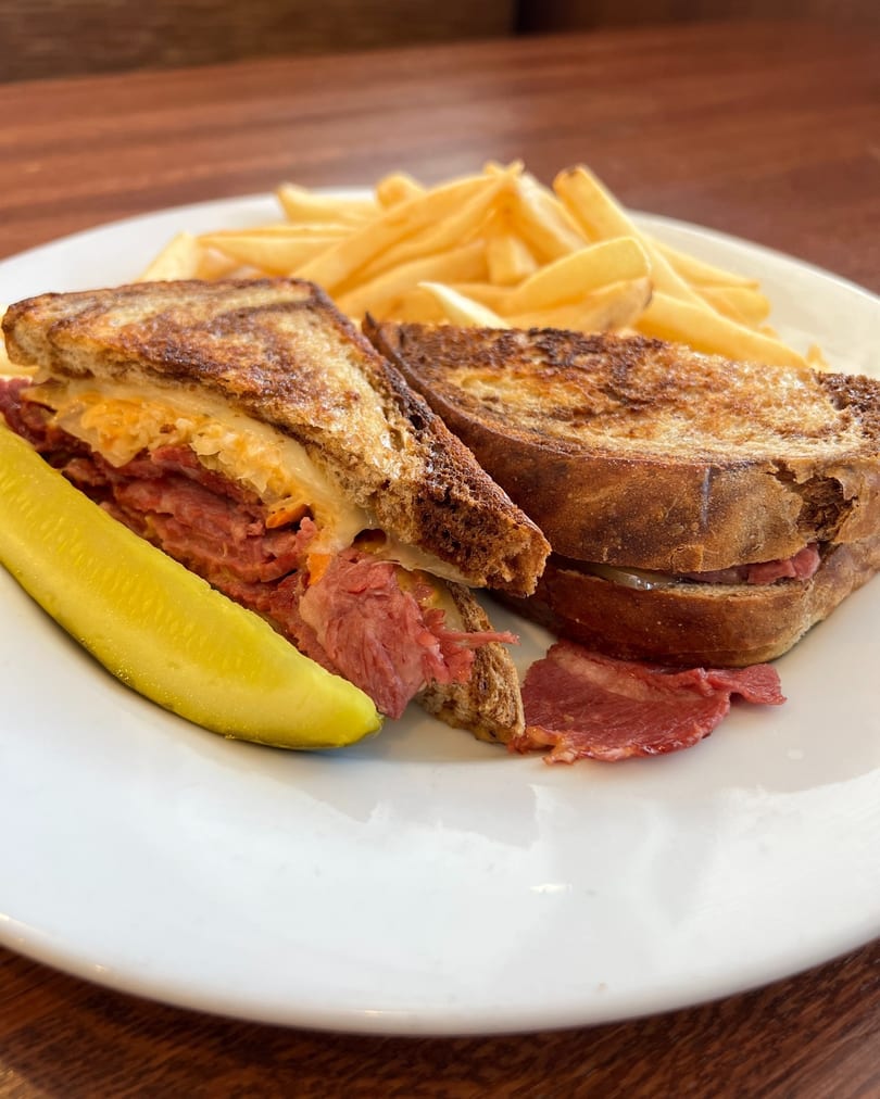 A grilled Reuben sandwich with corned beef, sauerkraut, and cheese, served with fries and a pickle spear.