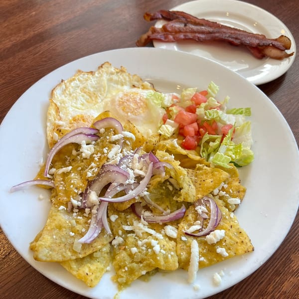 Chilaquiles with green sauce, fried eggs, red onions, cheese, lettuce, tomatoes, and bacon on the side.