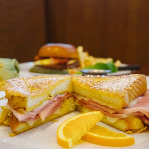 Close-up of a Monte Cristo sandwich with powdered sugar and orange slices, with other dishes blurred in the background.