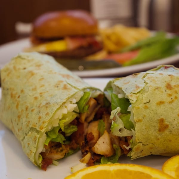A green tortilla wrap filled with chicken and lettuce, cut in half, with orange slices and a burger in the background.