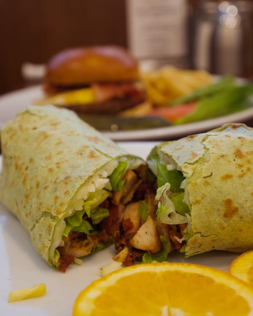 A green tortilla wrap filled with chicken and lettuce, cut in half, with orange slices and a burger in the background.